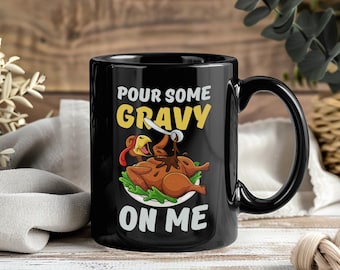 Può includere: Tazza in ceramica nera con un design umoristico del Ringraziamento. La tazza presenta il testo "Pour Some Gravy On Me" sopra un'illustrazione di un tacchino arrosto cosparso di salsa. La tazza ha un manico e un interno neri.