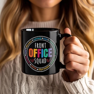 Puede incluir: Taza de cerámica negra con las palabras "FRONT OFFICE SQUAD" en letras coloridas. La taza tiene un asa negra y un diseño circular blanco con palabras como "confiable", "cariñoso" y "divertido".