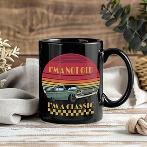 May include: Black ceramic mug with a retro design. The mug features a sunset graphic with a classic car and the text "I'M A NOT OLD I'M A CLASSIC". The mug has a black handle and a glossy finish.