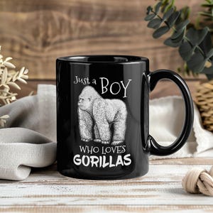 May include: Black ceramic mug with a white illustration of a gorilla and the text "Just a BOY who loves GORILLAS". The mug has a black handle and is set against a wooden background.