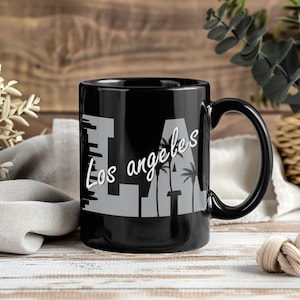 May include: Black ceramic mug with the text "LA Los Angeles" in white and gray. The design includes palm tree silhouettes. The mug has a black handle and is suitable for hot or cold beverages.
