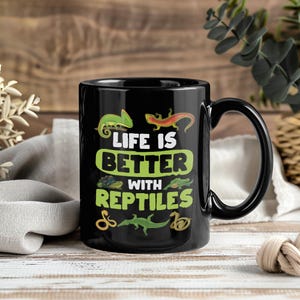 Op de afbeelding: Zwarte keramische mok met de tekst "LIFE IS BETTER WITH REPTILES" in wit en groen. De mok heeft illustraties van verschillende reptielen, waaronder een kameleon, een hagedis en een slang. De mok heeft een zwart handvat en een glanzende afwerking.