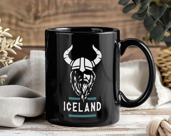 May include: Black ceramic mug with a white Viking helmet and beard design. The word "ICELAND" is printed below the graphic in white, with a light blue border. The mug has a black handle and is suitable for hot or cold drinks.
