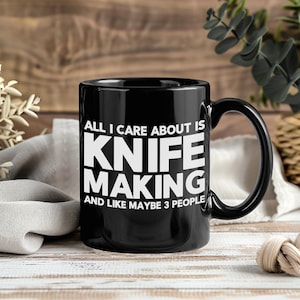May include: Black ceramic mug with the text "ALL I CARE ABOUT IS KNIFE MAKING AND LIKE MAYBE 3 PEOPLE" in white block letters. The mug has a large handle and is set against a wooden background.