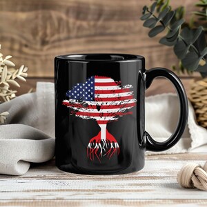 Puede incluir: Taza de cerámica negra con un diseño de árbol estilizado. La copa es la bandera estadounidense en rojo, blanco y azul. Las raíces también son rojas. La taza tiene un asa curva. Fondo de madera borroso.