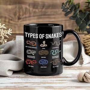 Puede incluir: Una taza de cerámica negra con el texto blanco TYPES OF SNAKES en la parte superior. Debajo del título hay nueve serpientes ilustradas en varios colores, cada una etiquetada con su nombre, incluyendo Grey-Banded Kingsnake, Cobra y Ball Python.