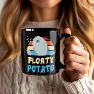Puede incluir: Taza de cerámica negra con una ilustración de foca de dibujos animados y el texto "FLOATY POTATO". La foca es de color gris claro, sobre un diseño de puesta de sol en rojo, naranja y azul.