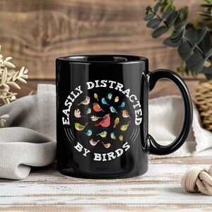 Könnte beinhalten: Schwarze Keramik-Tasse mit einem weißen, kreisförmigen Design mit verschiedenen bunten Vogelillustrationen. Der Text "EASILY DISTRACTED BY BIRDS" ist um das Design herum gedruckt. Die Tasse hat einen großen Henkel.