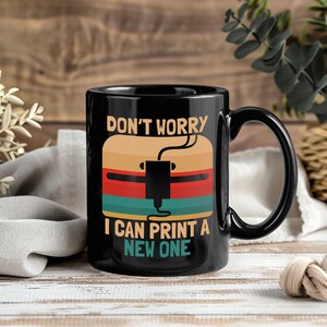 Könnte beinhalten: Schwarze Keramik-Tasse mit der Aufschrift "DON'T WORRY I CAN PRINT A NEW ONE". Die Grafik zeigt ein 3D-Drucker-Design mit Retro-Farbbalken in Beige, Rot und Türkis. Die Tasse hat einen schwarzen Henkel.