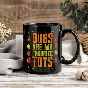 May include: Black ceramic mug with the text "BUGS ARE MY FAVORITE TOYS" in orange and yellow. The mug features illustrations of various bugs. The mug has a black handle and is set against a wooden background.