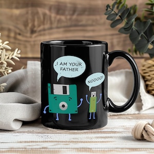 Op de afbeelding: Zwarte keramische mok met een humoristisch ontwerp. Een groen floppy disk-personage zegt "I AM YOUR FATHER" tegen een groen USB-stick-personage dat antwoordt "NOOOO!". De mok heeft een zwart handvat en een glanzende afwerking.