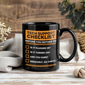 Puede incluir: Taza de cerámica negra con el texto "TECH SUPPORT CHECKLIST" en naranja y blanco. La lista de verificación incluye preguntas como "¿ESTÁ CONECTADO?" y "¿ESTÁ SEGURO?" Un regalo humorístico.
