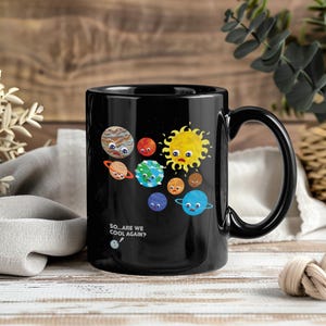 Peut inclure: Mug en céramique noire avec une illustration de dessins animés de planètes et du soleil avec des visages. La tasse a une anse noire et le texte "SO...ARE WE COOL AGAIN?" est imprimé sur le côté.