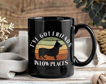 May include: Black ceramic mug with a wraparound handle. The mug features a graphic of a black corgi silhouette inside a retro-style sunset circle. Text reads "I'VE GOT FRIENDS IN LOW PLACES."