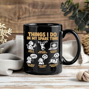 Può includere: Una tazza in ceramica nera con il testo "THINGS I DO IN MY SPARE TIME" in oro, accompagnata da sei illustrazioni grafiche bianche di scimmie con le didascalie: "LOOK AT MONKEYS", "HANG OUT WITH MONKEYS", "RESEARCH MONKEYS", "TALK ABOUT MONKEYS", "THINK ABOUT MONKEYS" e "DREAM ABOUT MONKEYS".