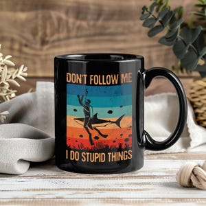 Peut inclure: Mug en céramique noire avec un design humoristique. L'illustration représente un plongeur et un requin avec le texte "DON'T FOLLOW ME I DO STUPID THINGS" en orange et blanc. Le fond présente des rayures horizontales bleues, oranges et turquoise.