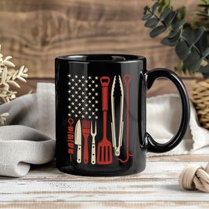 Peut inclure: Un mug en céramique noire orné d'un motif représentant un drapeau composé d'étoiles blanches et d'ustensiles de barbecue rouges. Les outils illustrés incluent une brochette, un couteau, un pinceau, une fourchette, une spatule, une pince et un crochet.
