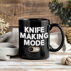 May include: A black ceramic mug with the words "KNIFE MAKING MODE" in white, bold letters. Below the text is a graphic of an "ON" switch. The mug has a black handle and is set against a wooden background.