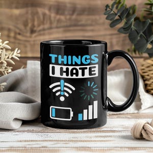 May include: Black ceramic mug with the text "THINGS I HATE" in white and blue. The mug features white and blue graphics of a wifi signal, a loading symbol, a low battery, and a signal bar.