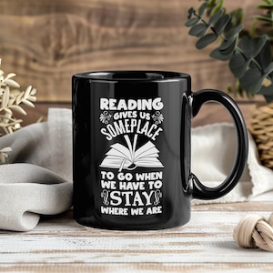 May include: Black ceramic mug with a white graphic that reads "Reading gives us someplace to go when we have to stay where we are." The mug has a large handle and is set against a blurred wooden background.