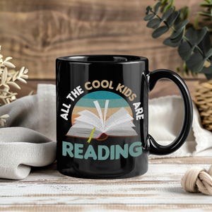 May include: Black ceramic mug with the text "ALL THE COOL KIDS ARE READING" in white, teal, and orange. The design includes an open book illustration. The mug has a black handle and is set against a wooden background.