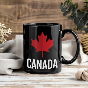 May include: A black ceramic mug featuring a prominent red maple leaf graphic centered on the front. Below the maple leaf, the word CANADA is printed in bold, white, sans-serif capital letters. The mug has a smooth, glossy finish and a handle.