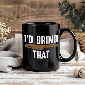 May include: Black ceramic coffee mug with the text "I'D GRIND THAT" in white, featuring a brown knife graphic. The mug has a black handle and is suitable for hot or cold beverages.