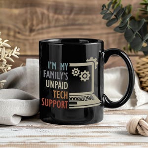 May include: Black ceramic mug with the text "I'M MY FAMILY'S UNPAID TECH SUPPORT" in white, beige, and red. The mug features a laptop graphic with gears. The mug has a black handle and is on a wooden surface.