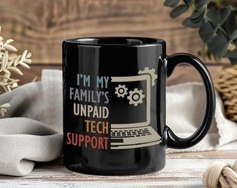 May include: Black ceramic mug with the text "I'M MY FAMILY'S UNPAID TECH SUPPORT" in white, beige, and red. The mug features a laptop graphic with gears. The mug has a black handle and is on a wooden surface.