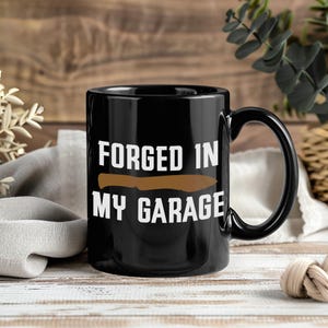 May include: Black ceramic mug with a large handle. The mug features the text "FORGED IN MY GARAGE" in white, with a brown knife graphic in the center. The mug is on a wooden surface.