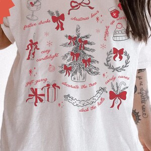 May include: White t-shirt with a festive Christmas design. The graphic includes a Christmas tree, bows, gift boxes, and text like "festive drinks" and "deck the halls" in red. The shirt has a relaxed fit and features the text "REBELBABE ORIGINAL DESIGN".