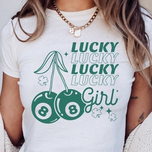 Könnte beinhalten: Ein weißes Crop-Top mit einem grünen Kirsch-Design. Die Kirsche hat zwei 8-Ball-Designs und der Stiel hat Kleeblätter. Der Text "Lucky Lucky Lucky Lucky Girl" ist in einer grünen Retro-Schrift geschrieben.
