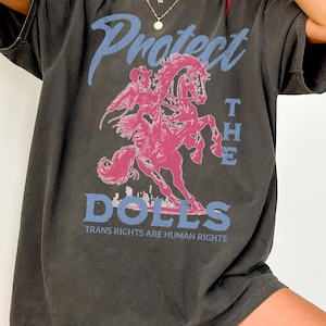 Puede incluir: Camiseta gris oscuro con un gráfico rosa y azul. El gráfico presenta una persona montando a caballo con las palabras "Protect the Dolls" y "Trans Rights are Human Rights". La camiseta tiene mangas cortas y un corte holgado.