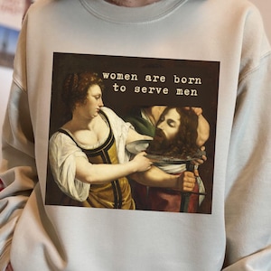 Può includere: Una felpa beige con un dipinto di una donna che tiene un piatto con la testa mozzata di un uomo. Il testo "women are born to serve men" è stampato sopra il dipinto.