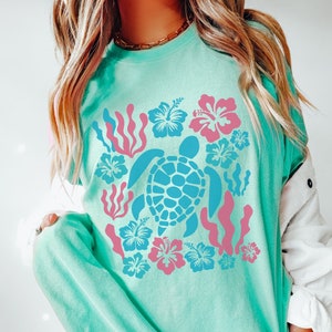 Peut inclure: Un t-shirt bleu turquoise avec un motif de tortue de mer entourée de fleurs d'hibiscus roses et bleues et d'algues.