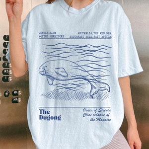 May include: Light blue t-shirt with a drawing of a dugong swimming in the ocean. The text on the shirt reads "The Dugong" and "Order of Sirenia Close relative of the Manatee".
