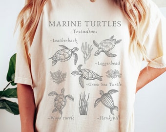 May include: Cream-colored t-shirt featuring a detailed illustration of various marine turtles, including Leatherback, Loggerhead, Green Sea Turtle, Wood turtle, and Hawksbill. The text "MARINE TURTLES Testudines" is printed at the top.