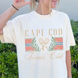 Puede incluir: Una camiseta blanca con un diseño gráfico de estilo vintage que presenta el texto "Cape Cod Tennis Club" con dos raquetas de tenis y una pelota de tenis. El gráfico está en tonos de rojo, azul, verde y dorado.