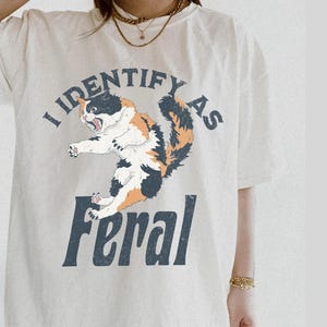 May include: Cream-colored t-shirt with a graphic of a calico cat mid-leap, mouth open in a roar. The text "I Identify as" arches above the cat, with "Feral" below in a bold, dark blue font.
