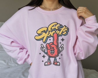 May include: Pink sweatshirt with a cartoon hand wearing a red glove and the text "Go Sports" in a retro style.