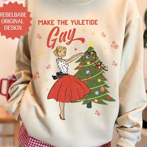 May include: Beige sweatshirt with a retro illustration of a woman decorating a Christmas tree. The text "MAKE THE YULETIDE GAY" is above the illustration. A red starburst logo reads "REBELBABE ORIGINAL DESIGN".