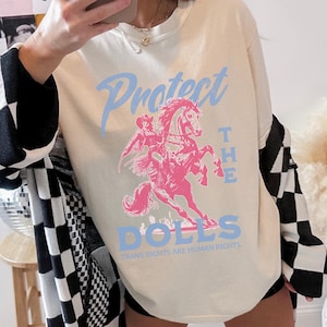 May include: Cream-colored sweatshirt with a pink graphic of a person riding a horse, and the words "Protect the Dolls" in light blue. The phrase "Trans Rights Are Human Rights" is printed below the graphic. The sweatshirt has long sleeves.