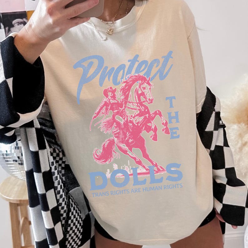 May include: Cream-colored sweatshirt with a pink graphic of a person riding a horse, and the words "Protect the Dolls" in light blue. The phrase "Trans Rights Are Human Rights" is printed below the graphic. The sweatshirt has long sleeves.