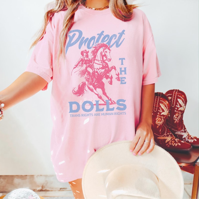 May include: Pink t-shirt with a graphic of a person riding a horse, and the words "Protect the Dolls" in blue. The text "Trans Rights Are Human Rights" is printed below the graphic. A pair of red cowboy boots are visible in the background.