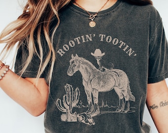 Op de afbeelding: Een donkergrijs T-shirt met een vintage-stijl afbeelding van een cowboy die op een paard rijdt met de tekst "ROOTIN' TOOTIN'". De afbeelding bevat ook een cactus en een schedel.