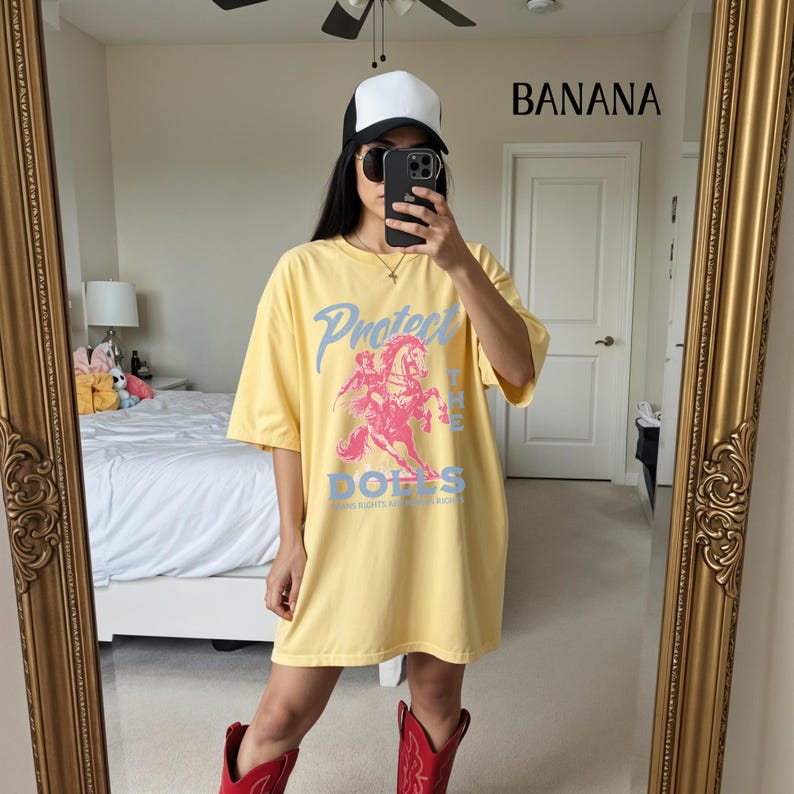 May include: A yellow t-shirt with the text "Protect the Dolls" and a graphic of a person riding a horse. The shirt is paired with red cowboy boots and a white and black baseball cap.