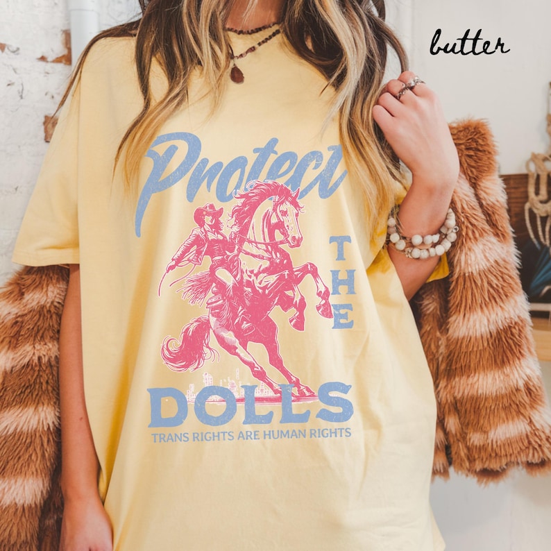 May include: Butter-colored t-shirt with a pink and blue graphic. The graphic features a cowboy riding a horse, with the words "Protect The Dolls" and "Trans Rights Are Human Rights" in blue text. The word "butter" is written in cursive.