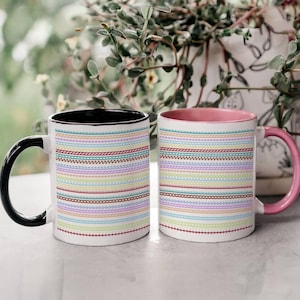 May include: Two white ceramic mugs with colorful horizontal stitch patterns. One mug has a black handle and interior, the other has a pink handle and interior. The mugs are decorated with rows of various stitches in pastel colors.