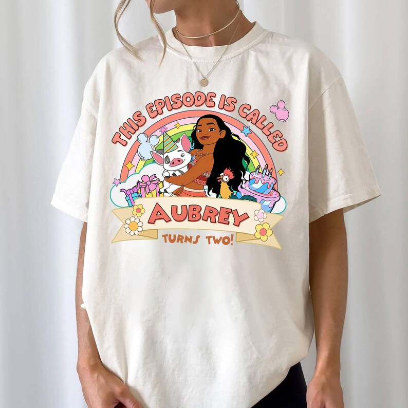 May include: Cream-colored t-shirt featuring a colorful graphic with the text "THIS EPISODE IS CALLED" above a rainbow. Below the rainbow is the name "AUBREY" and the phrase "TURNS TWO!". The graphic includes cartoon characters, a cake, and flowers.