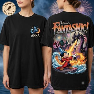 May include: A black t-shirt featuring a small graphic on the front with a wizard hat and the name Anna. The back displays a large, colorful Fantasmic graphic with Mickey Mouse in a wizard robe, a dragon, water, fire, and fireworks in the night sky.